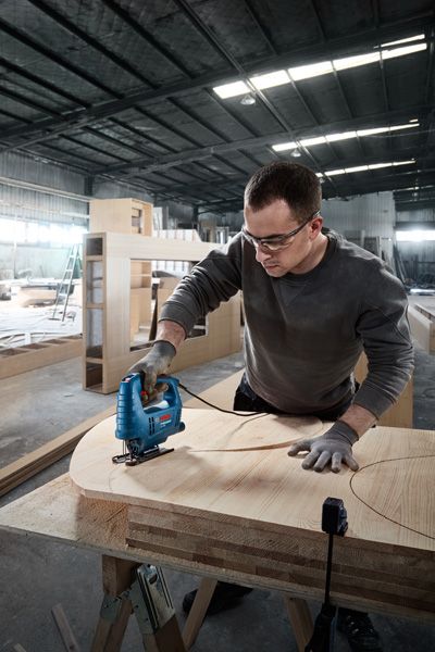 Person wearing safety equipment uses a jigsaw to cut curved shapes in plywood.