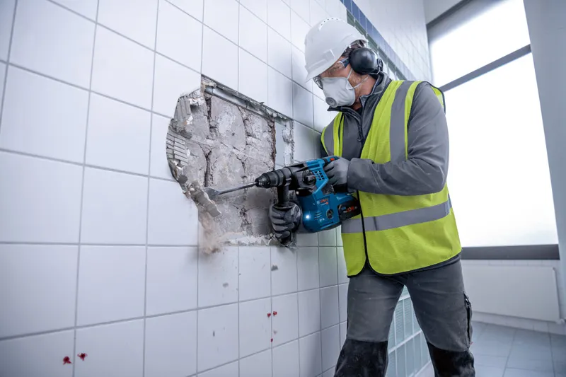 Person wearing safety equipment uses a cordless rotary hammer to break through a tiled wall.