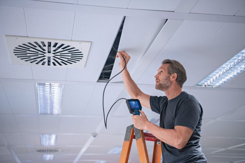 A person stands on a ladder using an inspection camera to examine a ceiling panel.