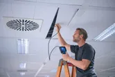 A person stands on a ladder using an inspection camera to examine a ceiling panel.