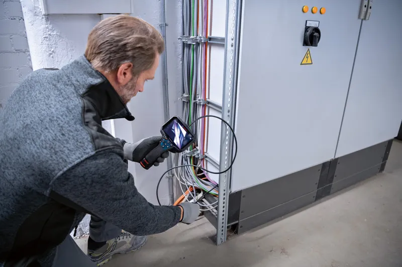 A person wearing safety equipment inspects electrical wiring with an inspection camera.