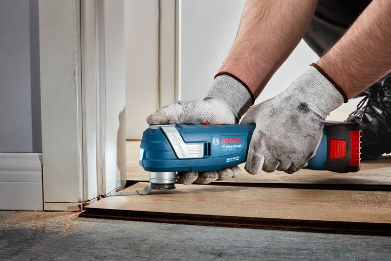 A person wearing safety equipment trims wood flooring with a cordless multi-tool.