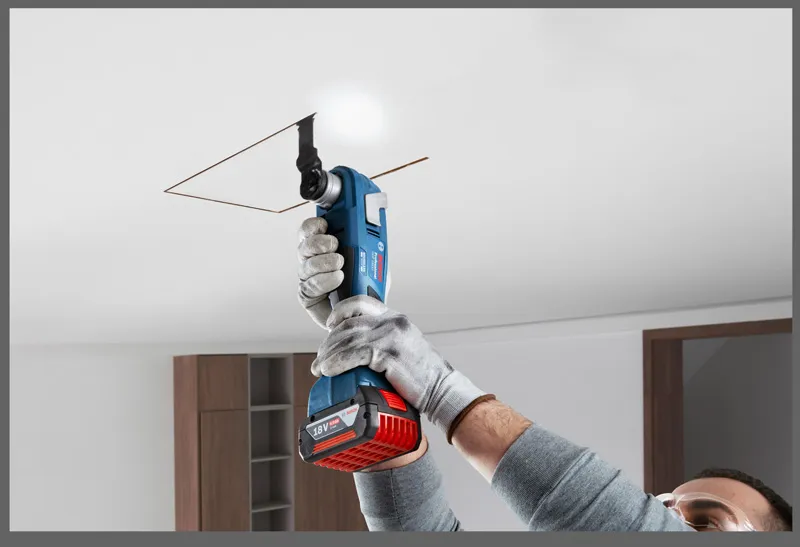 A person wearing safety equipment cuts a ceiling panel with a cordless oscillating tool.