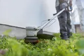 一名配戴安全設備的人員正在使用無線割草機沿牆修剪草坪。.