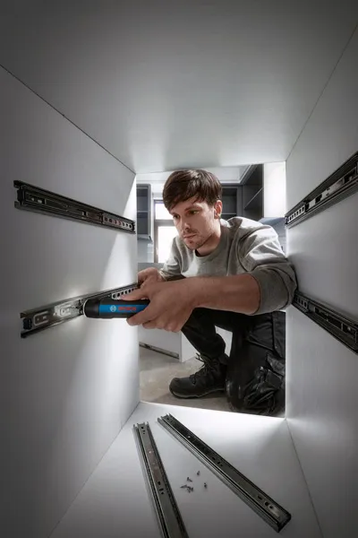 A person uses a cordless screwdriver to install drawer slides inside a cabinet.