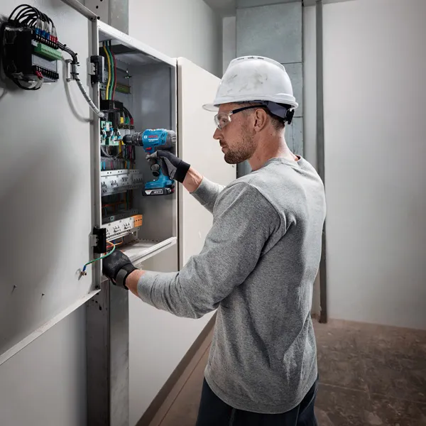 一名佩戴安全設備的人員使用無線電鑽在電氣面板上工作。.