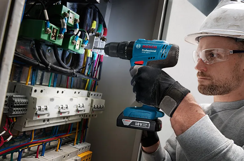 一名佩戴安全設備的人正在鑽進佈滿電線的控制面板。.