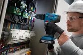 一名配戴安全裝備的人員使用電鑽在配電盤上作業。