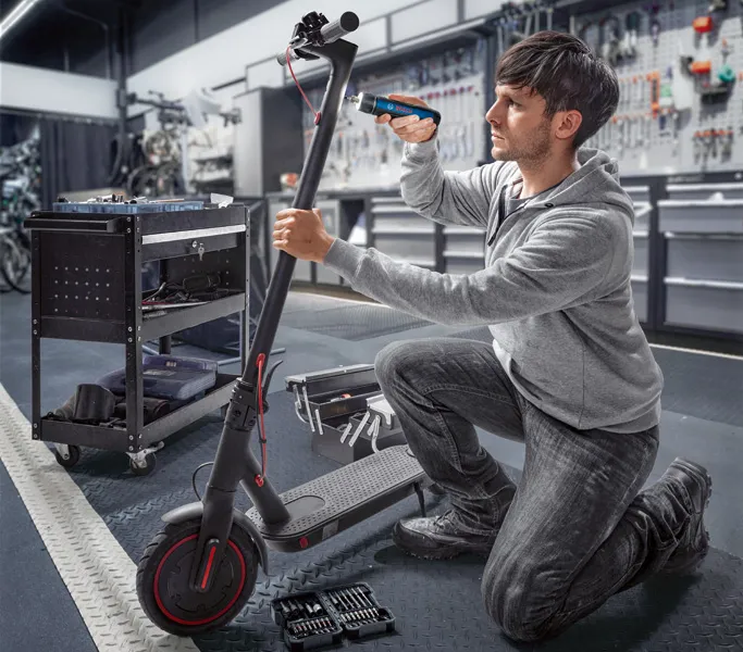 A person repairs an electric scooter using a cordless screwdriver in a workshop.