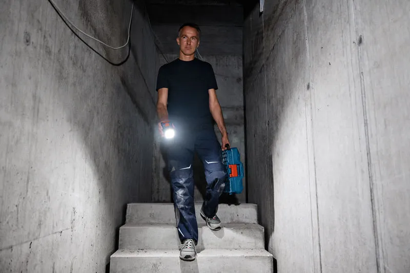 A person wearing safety equipment walks down concrete stairs holding a flashlight and a tool case.