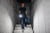 A person wearing safety equipment walks down concrete stairs holding a flashlight and a tool case.