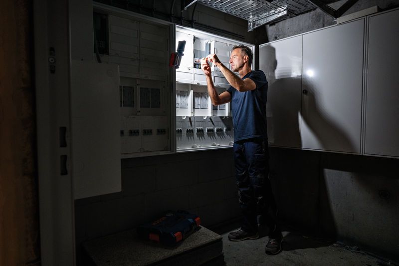 A person wearing safety equipment repairs an electrical panel under bright task lighting.