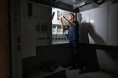 A person wearing safety equipment repairs an electrical panel under bright task lighting.