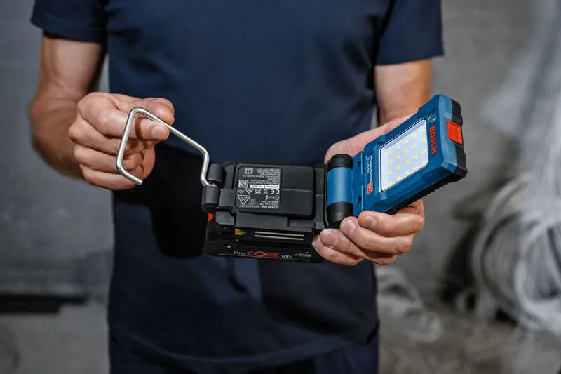 Person holds a compact LED work light with attached battery in an industrial setting.
