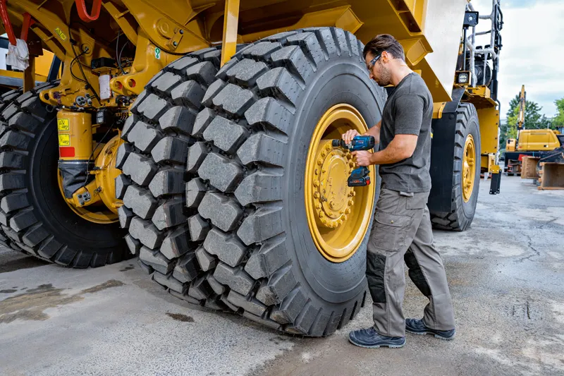 一名配戴安全設備的人員使用衝擊扳手來擰緊大型工程車輛的輪胎。.