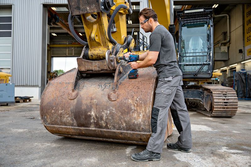 A person wearing safety equipment uses an impact wrench on heavy construction machinery.