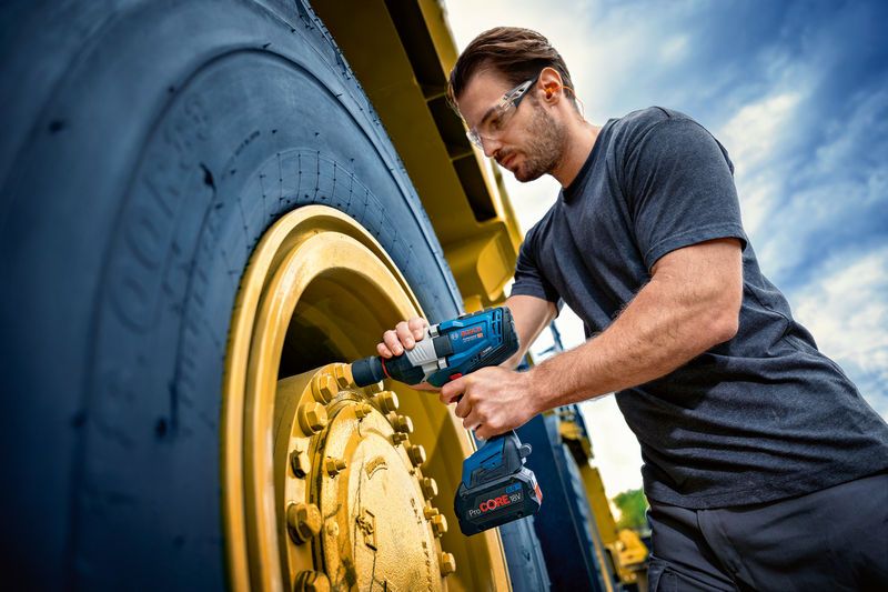 A person wearing safety equipment uses an impact wrench on a large vehicle wheel.