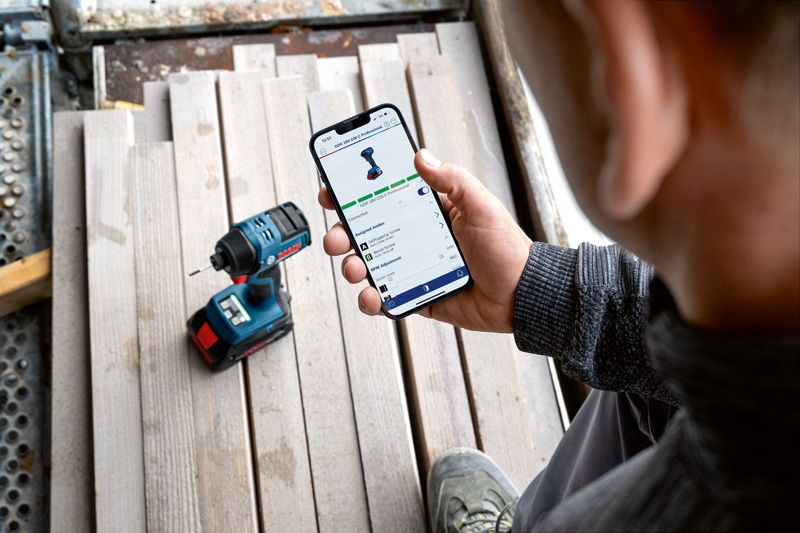 A person checks a power tool's app features on a smartphone near a cordless drill.