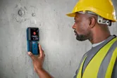A person wearing safety equipment uses a detector on a concrete wall.