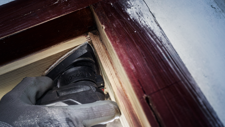 一名身穿安全設備的人員在油漆過的窗框附近打磨木材。