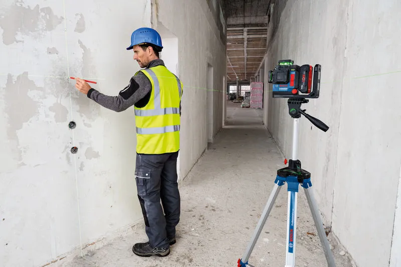 Worker wearing safety equipment marks a wall using a laser leveling tool on a tripod.