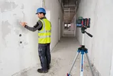 Worker wearing safety equipment marks a wall using a laser leveling tool on a tripod.