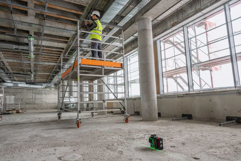 A person wearing safety equipment stands on scaffolding while using a laser leveling tool.