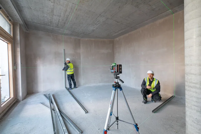 Two workers wearing safety equipment align metal studs using a laser leveling tool in an unfinished room.