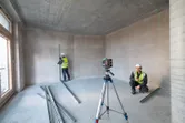 Two workers wearing safety equipment align metal studs using a laser leveling tool in an unfinished room.