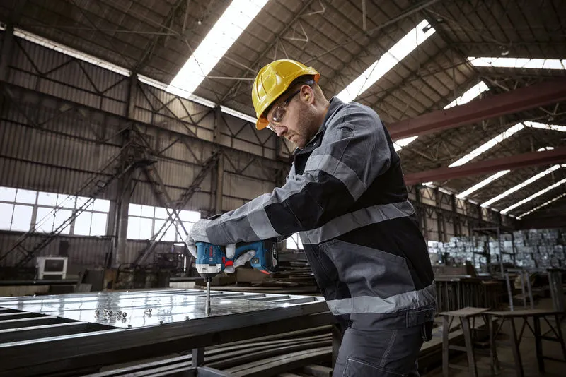 一名配戴安全設備的工人在工廠裡使用電鑽鑽打金屬框架。.