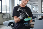 一名配戴安全設備的人員在汽車修理廠內對無線拋光機進行拋光。.