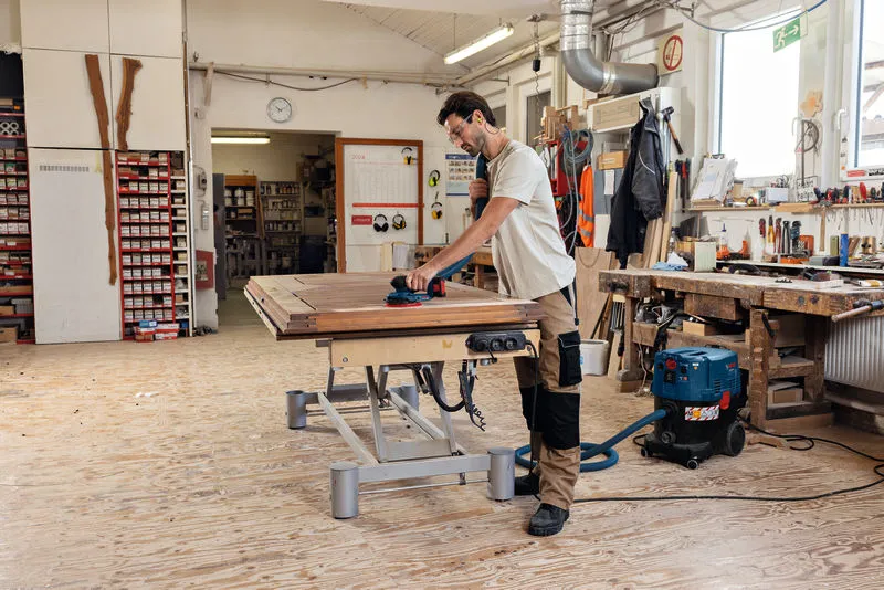 一名配戴安全設備的人員正在使用無線隨機軌道砂光機打磨木質表面。.