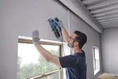 一名配戴安全設備的人員在窗戶附近使用軌道式砂光機打磨牆壁。.