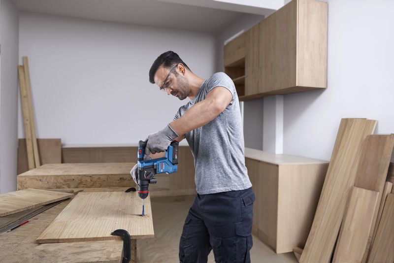 一名佩戴安全設備的人使用無線旋轉錘在木頭上鑽孔。.