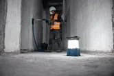 一名穿著安全設備的工人正在牆上鑽孔，無線工地燈放在地板上。.