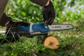 A person wearing safety equipment adjusts a cordless pole saw on a cut log outdoors.