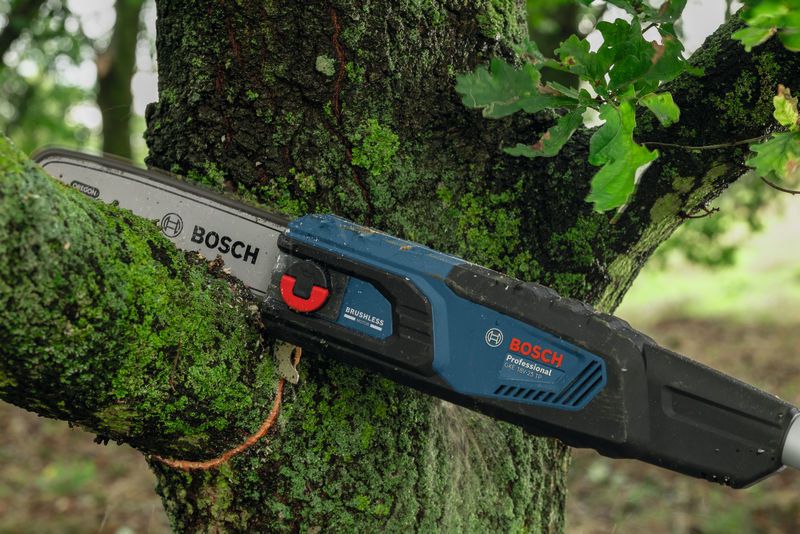 Cordless pole saw cutting through a mossy tree branch outdoors.