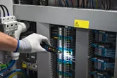 一名配戴安全設備的人員正在使用電壓測試器檢查控制面板中的電線。