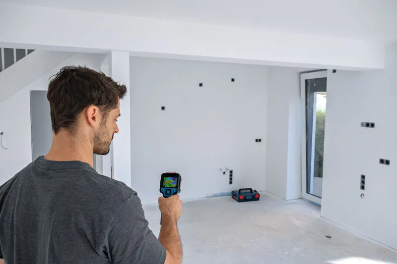 A person uses a thermo camera to scan a wall in an unfinished room.
