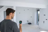 A person uses a thermo camera to scan a wall in an unfinished room.