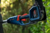 A person wearing safety equipment holds a cordless pole saw outdoors.