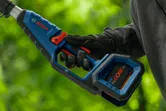 Person wearing safety equipment holds a cordless pole saw outdoors.