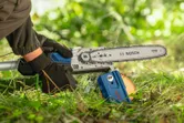 A person wearing safety equipment trims a log with a cordless pole saw outdoors.
