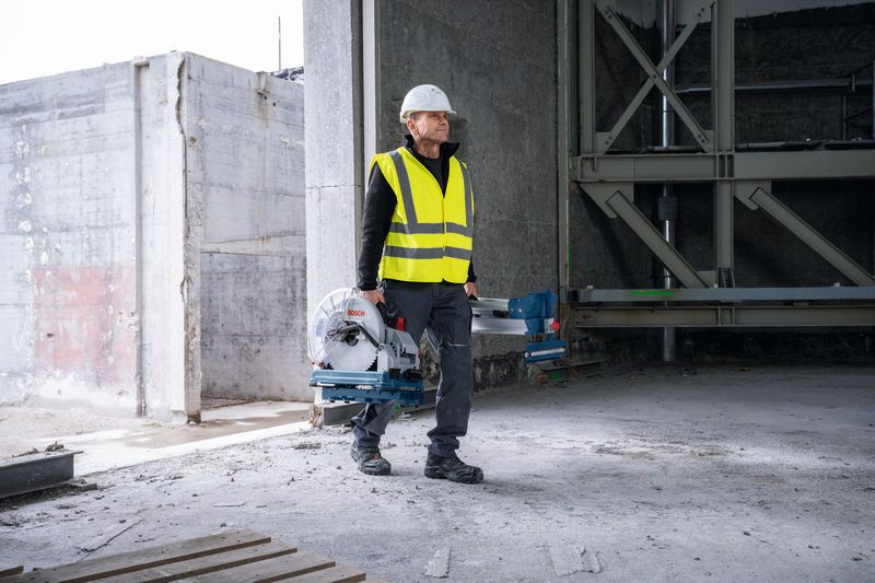 A person wearing safety equipment carries a metal cut-off saw in an unfinished building.