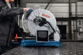 A person wearing safety equipment operates a metal cut-off saw on a steel pipe.