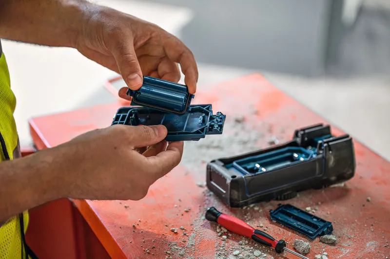 A person wearing safety equipment changes batteries in a power tool on a dusty workbench.