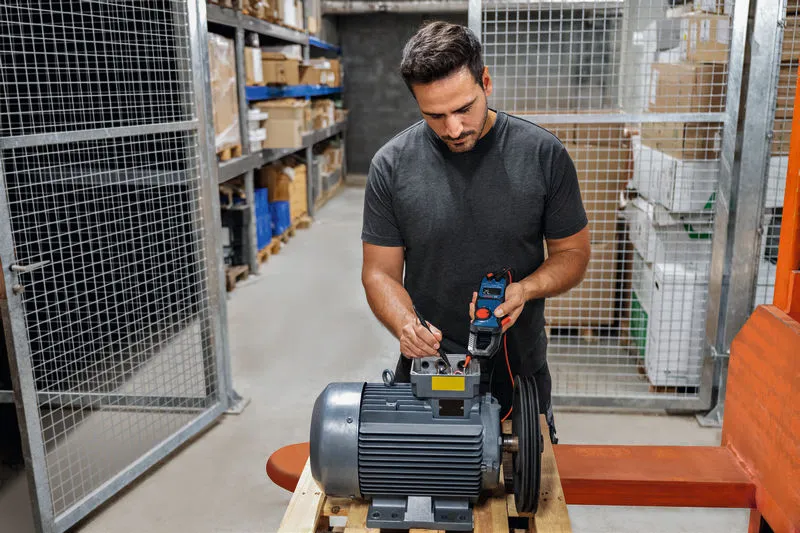 Person testing an industrial motor with a clamp meter in a warehouse.