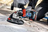 A person wearing safety equipment uses a cordless concrete grinder on a floor surface.