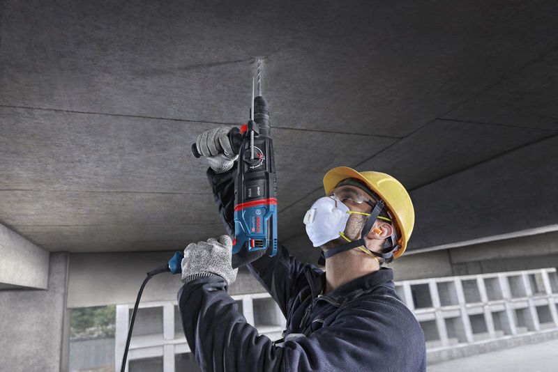 A person wearing safety equipment drills into a concrete ceiling using a rotary hammer.