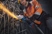 A worker wearing safety equipment uses an angle grinder to cut metal rebar.